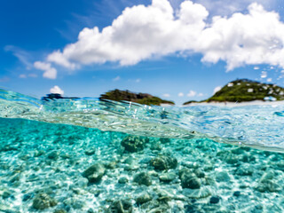 嘉比島の白砂ビーチの半水面撮影。沖縄特有の双胴船サバニや観光客、パラソル、テントが見える。美しいサンゴ礁と熱帯魚の群れ。 安室島沖縄県 島尻郡慶良間諸島座間味島-2025 サンゴの産卵があり卵が漂う。沖縄本島から40km西方の諸島。30以上の島嶼群全体が国立公園に指定。珊瑚礁の青い海と白い砂浜は世界屈指の美。貴重な動植物が多数生息。 