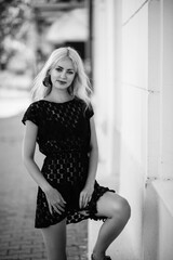 Young woman with long blonde hair in a black dress poses elegantly against a white wall, showcasing her fashion sense and confidence in a stylish urban setting