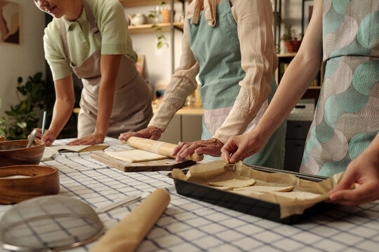 Three Caucasian women preparing dough together in kitchen, one rolling dough with rolling pin while others arranging pastry on baking tray, collaborating on homemade baking project