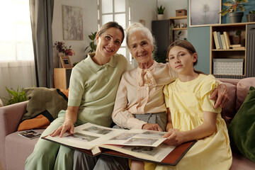 Caucasian middle aged woman, senior Caucasian woman, and Caucasian girl sitting on sofa looking at photo album together, smiling and embracing, family bonding moment