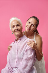 Caucasian senior woman sitting with Caucasian young adult woman standing behind, both smiling and looking at camera, showing affectionate family relationship against pink background