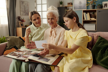 Senior Caucasian woman sitting on sofa with young adult Caucasian woman and preteen Caucasian girl, sharing memories while looking through photo album and holding photograph together
