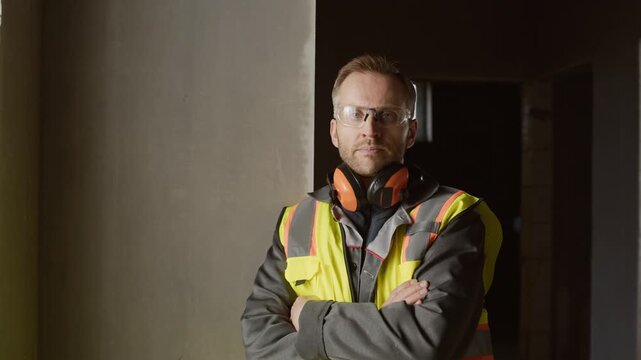 Portrait of confident construction professional in safety vest, earmuffs and protective glasses posing with folded arms at modern unfinished construction site