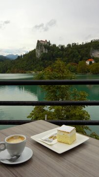 Coffee And Cream Cake At Cafe Overlooking The Lake Bled With Bled Castle In Slovenia. - vertical shot