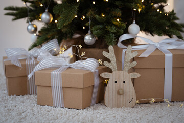 A wooden reindeer decoration with gold glitter details and Christmas present with white ribbon under a Christmas tree with lights in the background. Christmas decorations. Christmas theme.