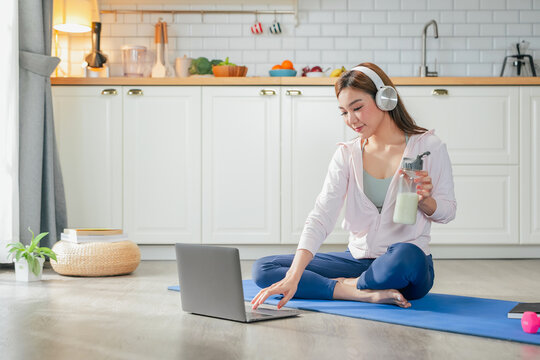 happy motivated Asian woman practices yoga at home online private class stretching body perfect balance listening instructor headphones holds protein shake embracing healthy lifestyle peaceful home