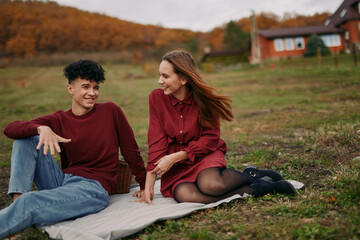 Fototapeta premium A young couple sharing a genuine outdoor picnic on a blanket near a rustic cottage, candid smiles and authentic interaction captured in warm autumn light, conveying real emotion and close connection.