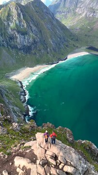 Hikers admire stunning vistas from the Ryten Kvalvika Beach Trail in Lofoten, Norway. The dramatic landscape features mountains, ocean, and sandy beach under a clear sky.