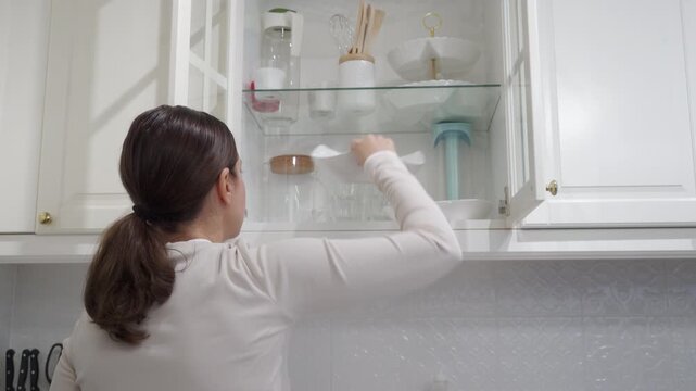 Woman Places Clean White Bowl in Open Overhead Glass-Front Kitchen Cabinet