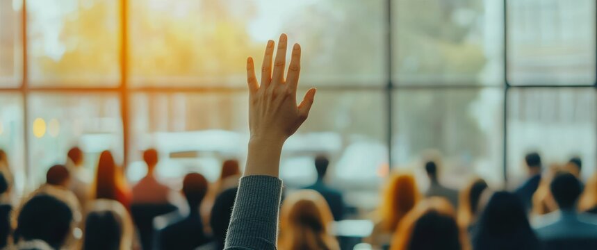 A person raising hand in a seminar or classroom setting with a blurred audience in the background and warm natural light coming through large windows