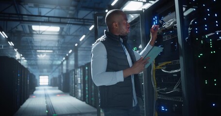 Data center technician uses a tablet to monitor and troubleshoot server rack hardware, checks network cables, inspects equipment in facility filled with organized cabling and rows of servers. - Powered by Adobe