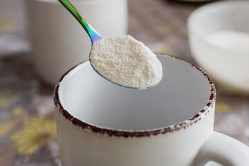 Tea spoon with dry powdered milk, closeup photo. Powdered milk in spoon over enamel mug, closeup on rustic kitchen table