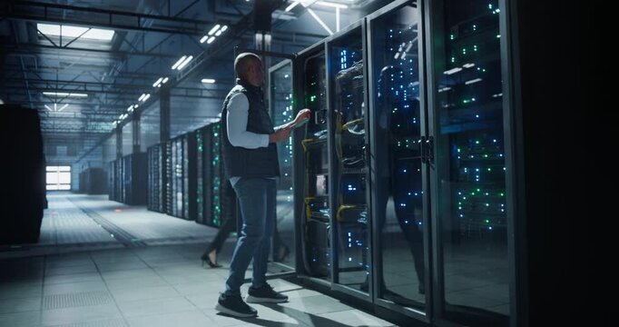 Data center IT specialist walks through aisles of server racks, using a tablet to inspect equipment, monitor network activity, and check connections in a cyber secure server room