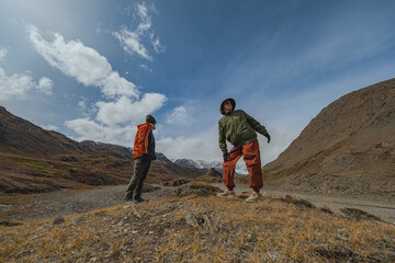 Fototapeta premium Two joyful hikers in bright clothes strike a funny pose on an autumn mountain trail with stunning snow-capped peaks in the background.