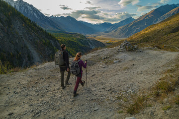 Two hikers with backpacks and trekking poles walk a narrow mountain trail alongside a steep cliff with a flowing river. A vast valley with autumn forests and distant mountains unfolds before them.