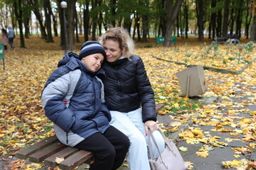 A woman and her son are sitting on a park bench and drinking tea with a croissant.