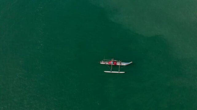 Breathtaking bird's eye view of a traditional outrigger canoe with fishermen gliding across calm emerald waters, minimalist tropical seascape perfect for travel and nature themes