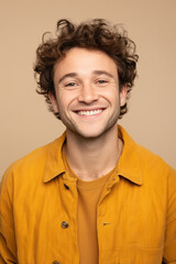 Obraz premium A young man with curly brown hair smiles at the camera. He wears a yellow collared shirt over a matching t-shirt. A neutral beige backdrop is featured.