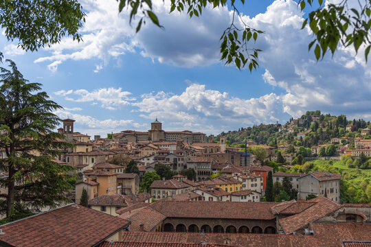 Panoramic view of Citta Alta in Bergamo reveals red roofs, church towers, and hillside villas, where historic center unfolds under a bright blue sky with drifting clouds