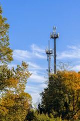 Vertical view of telecom towers above autumn forest, metal antennas rising into vivid blue sky framed by colorful foliage and gentle white cloud patterns