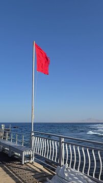 A red flag flutters in the wind on a pontoon by the sea. A strong wind and a red flag on the beach mean swimming is prohibited.