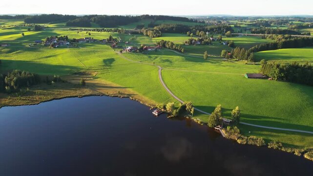 Aerial view of the dark waters of Attlesee lake meeting the bright green fields and scattered buildings under the sunlight, Attlesee, Bayern, Germany.