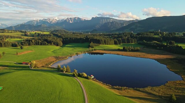 Aerial view of a serene lake reflecting the sky, surrounded by green fields, forests, and distant mountains, creating a tranquil landscape, Nesselwang, Bayern, Germany.