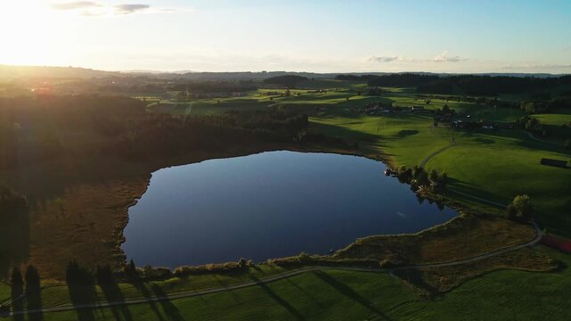 Aerial view of Attlesee lake reflecting the golden sunlight, surrounded by lush green fields and forests, creating a scenic landscape, Nesselwang, Bayern, Germany.