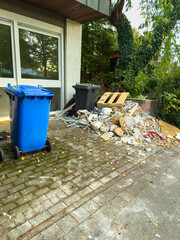 Fototapeta premium Construction debris accumulates beside blue and black waste bins in a quiet urban setting during late afternoon