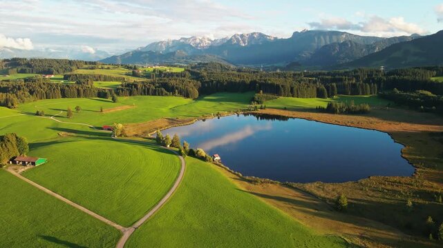 Aerial view of a serene lake mirroring the sky, surrounded by vibrant green fields and forests, creating a picturesque landscape, Nesselwang, Bayern, Germany.