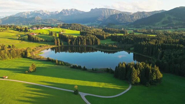 Aerial view of a picturesque lake surrounded by green fields, forests, and mountains offering a serene landscape, Nesselwang, Bayern, Germany.
