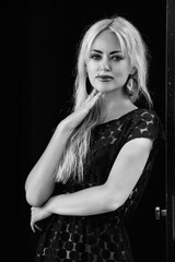 Elegant woman with long blonde hair, wearing a stylish black dress, poses confidently against a door, showcasing her fashion sense and strong presence in a monochrome setting