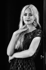 Elegant woman with long blonde hair, wearing a stylish black dress, poses confidently against a door, showcasing her fashion sense and strong presence in a monochrome setting