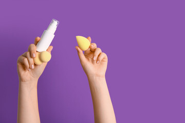 Female hands with makeup sponges and bottle of foundation on purple background
