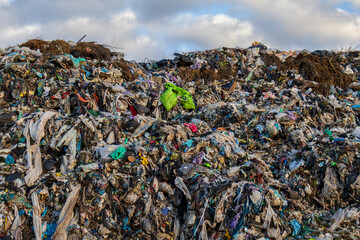 A large landfill site displays a chaotic mix of plastic waste, garbage, and organic matter under a cloudy sky, highlighting pollution issues