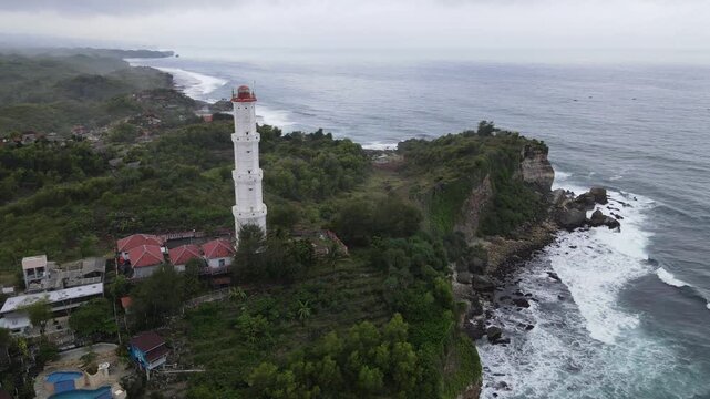 Aerial view of the Baron Lighthouse, a tall white structure on a cliff overlooking the ocean with crashing waves, Yogyakarta, Yogyakarta, Indonesia.