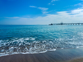 Serenity of the seashore at midday with waves gently lapping against the sandy beach and a distant pier extending into the calm blue sea