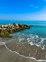 Fototapeta premium Beautiful coastline with gentle waves lapping against a rocky jetty under a clear blue sky at midday