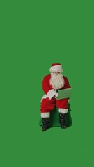 Santa Claus with a white curly beard, wearing a red suit and sunglasses, is working on a laptop while sitting on a green mockup against a green background.