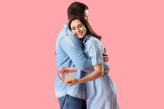 Happy young woman with gift hugging her husband on pink background