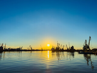 Golden sunset over bustling harbor with cranes and boats silhouetted against the colorful sky