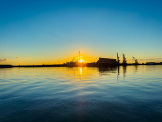 Fototapeta premium Golden sunset over calm waters reflecting silhouettes of boats and a distant shipyard in the evening sky