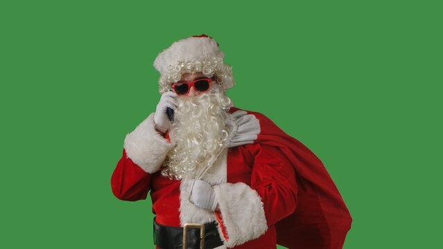 Santa Claus with a white curly beard, wearing a red suit and sunglasses, with a bag of gifts, talking on a smartphone against a green background. Perfect for Christmas themed compositions.