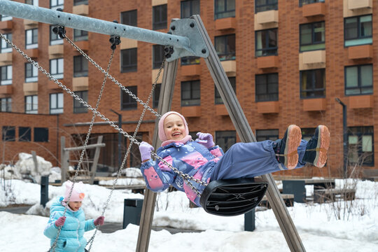 Happy little girls in snowy yard fun swing and laugh in winter playground. Children fun play walking outdoors.