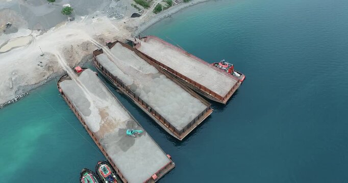Aerial view of barges laden with quarry aggregate, contrasting against the turquoise water and the pale earth of the quarry site, Palu, Central Sulawesi, Indonesia.