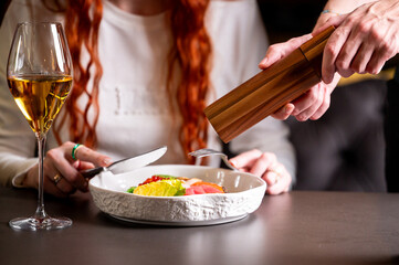A person is being served food in a restaurant, with a wooden pepper mill used for seasoning the colorful dish. A glass of white wine is nearby on the dark table