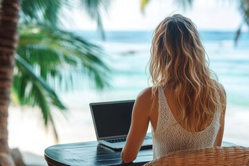Woman with long wavy hair wearing a white sleeveless top sitting at a round wooden table working on a laptop outdoors near a beach with palm trees and ocean in the background