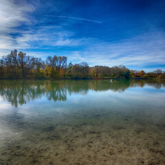 Germany, Bavaria - lake of Garching near Munich in a sunny day of autumn with the blue sky reflecting in the water and red leaves on the trees