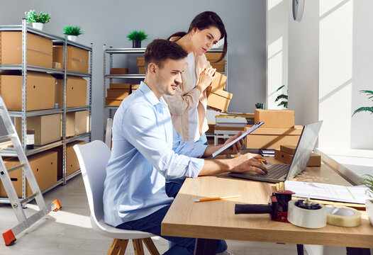 Young cheerful colleagues standing at workplace in storeroom checking stock and inventory on laptop. Company employees preparing order in storeroom in retail warehouse full of cardboard boxes.