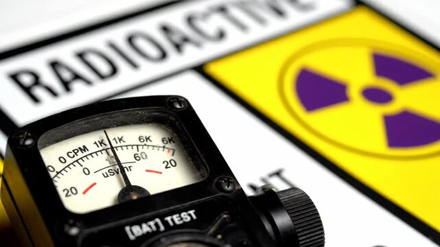 A close-up featuring a radioactive meter positioned above a bright yellow warning sign. The sign displays a purple trefoil symbol and stark black text, indicating radioactive material.